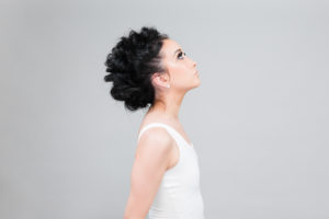 Profile of dark-haired woman with an edgy updo wearing a white tank top against a light gray backdrop