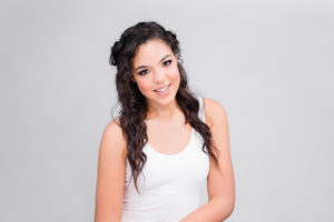 Model against neutral gray backdrop wearing a white tank top with dark hair. 