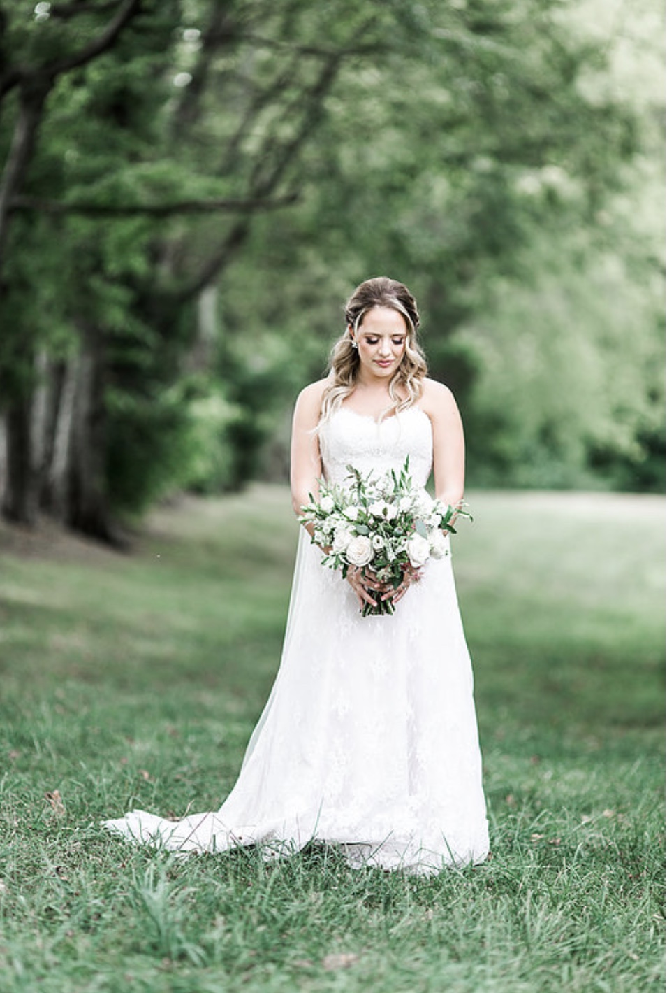 Bride standing in a green meadow looking toward the ground holding bouquet